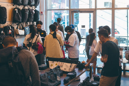 Customers browsing bags and SAMPLE pouches around the table at a UrbanCred meetup, with the West St windows behind